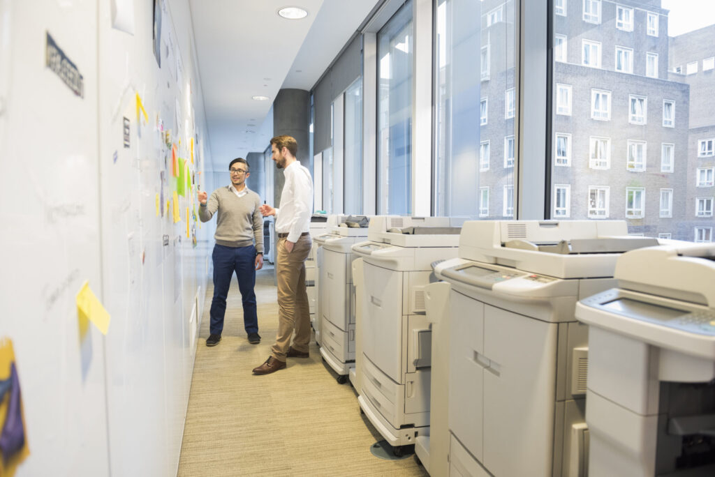 A full length photo of mid adult businessmen discussing over notes stuck on wall. Professionals are standing by computer printers. Copy machines are found near windows, in a brightly lit modern office alley.